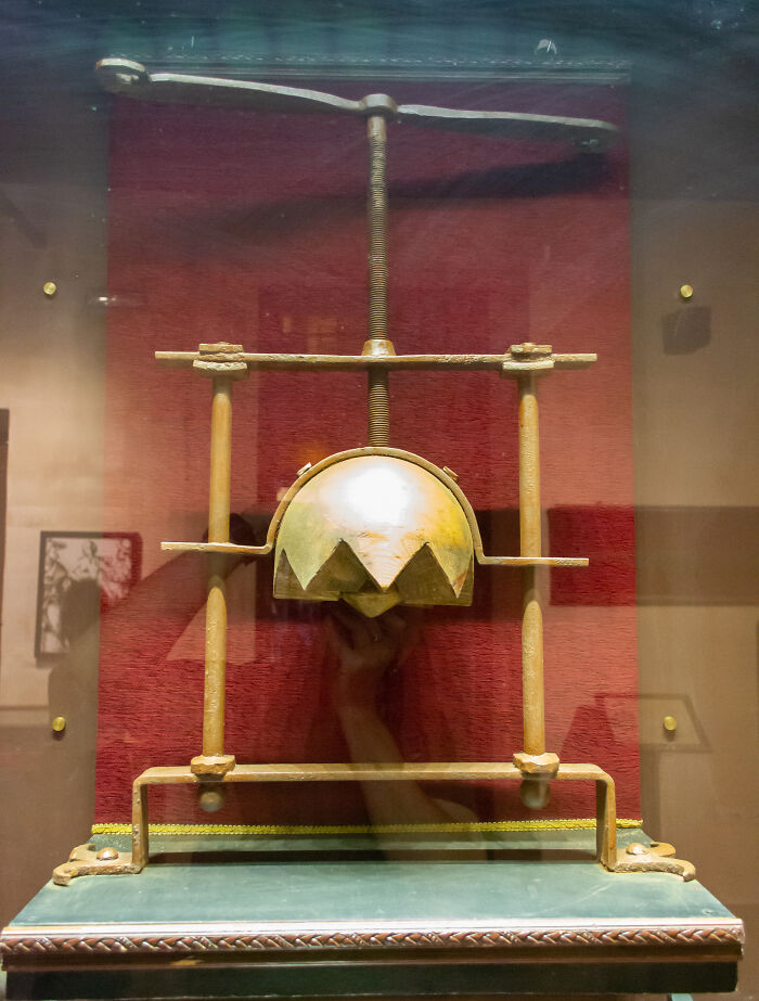 Medieval history t*****e device with iron spikes and s***w mechanism displayed on a red background in a museum.